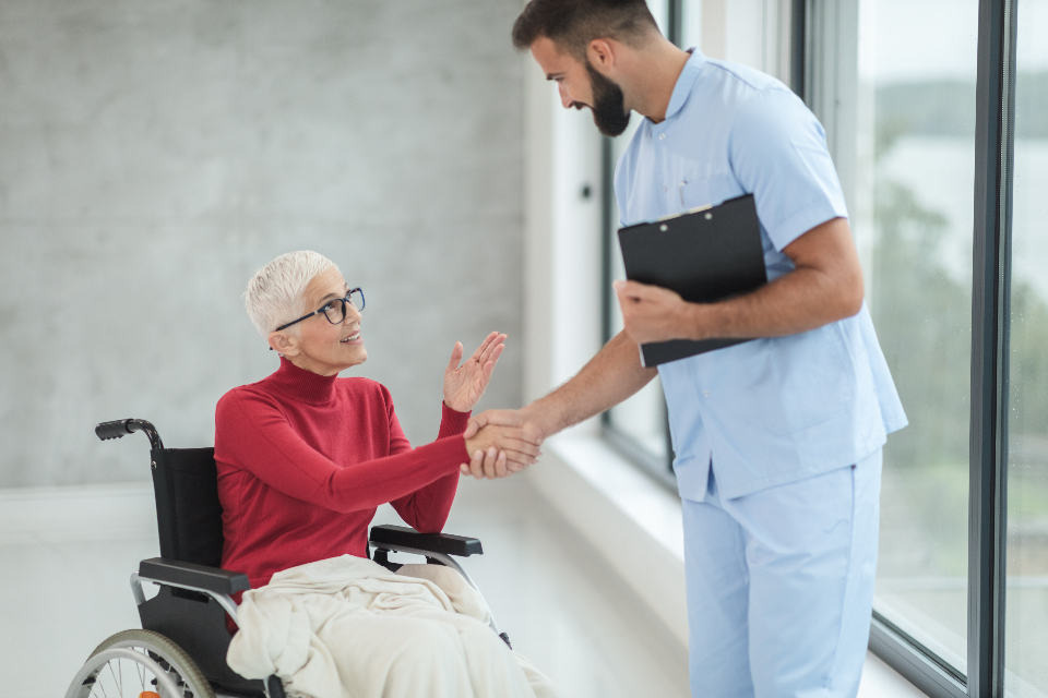 A warm caregiver in scrubs shaking hands with a smiling person who uses a wheelchair