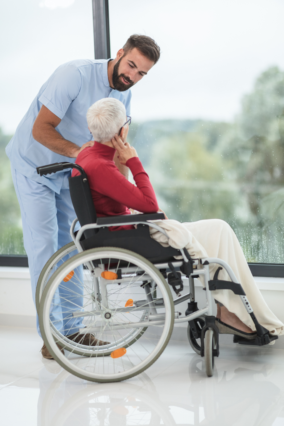 A smiling caregiver in scrubs gently supporting a person who uses a wheelchair by a bright window